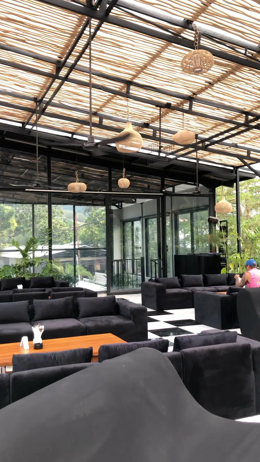 Lounge lantai atas OZ: sofa gelap dan meja kayu di bawah atap bambu, dikelilingi tanaman tropis.