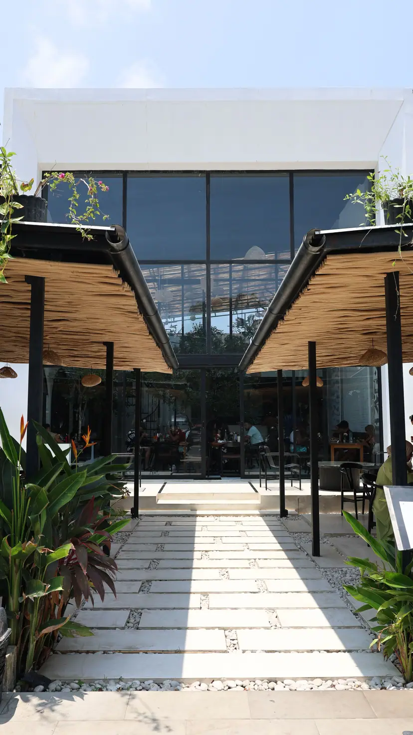 The entrance of OZ in daylight: a stone path between two woven roofs to the glass front.