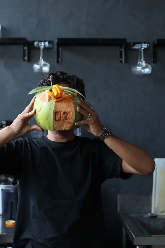 Un barman tient une noix de coco au logo OZ devant son visage.