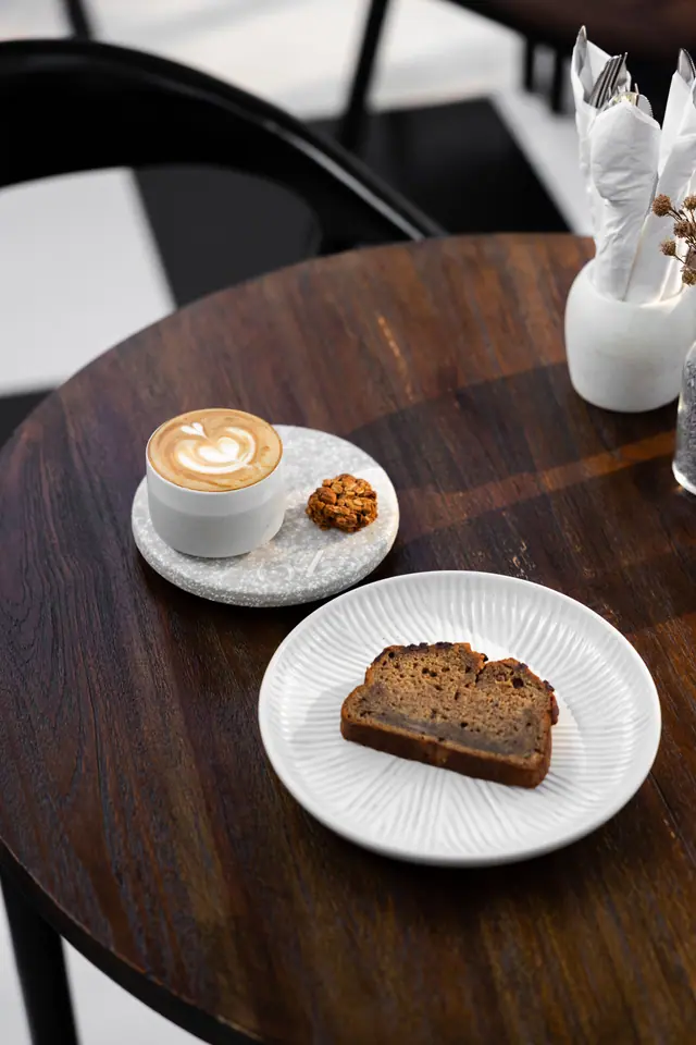 Un flat white et une tranche de banana bread sur une table ronde en bois.