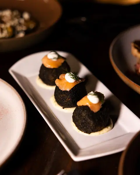 Three black arancini topped with smoked salmon on a white plate.