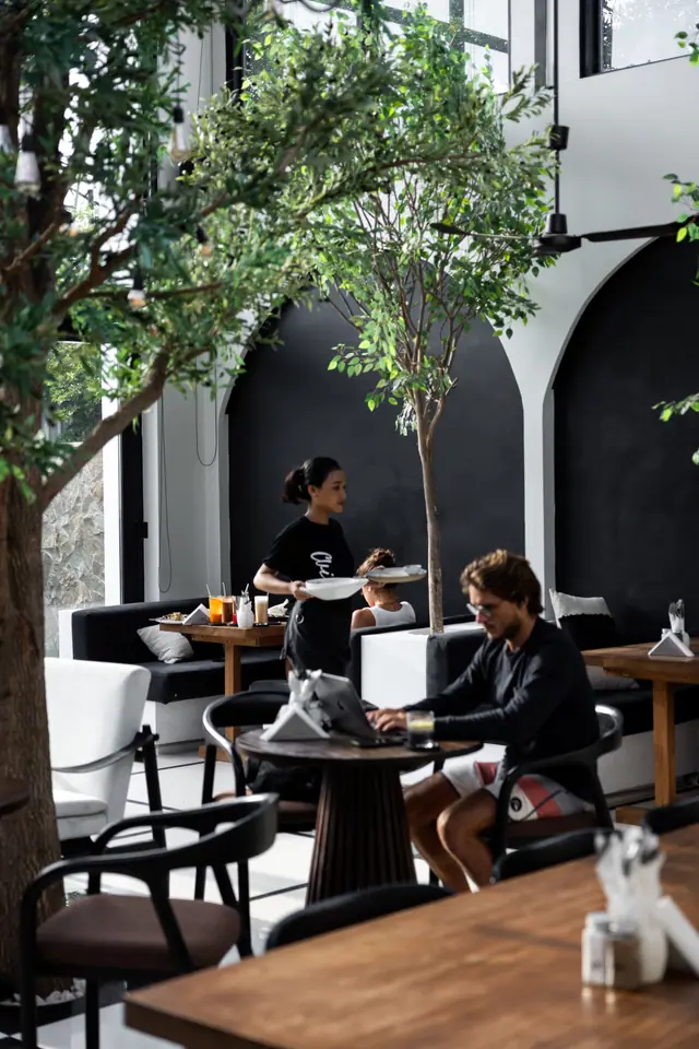 La grande salle blanche aux arches noires et aux arbres. Une serveuse apporte des assiettes.
