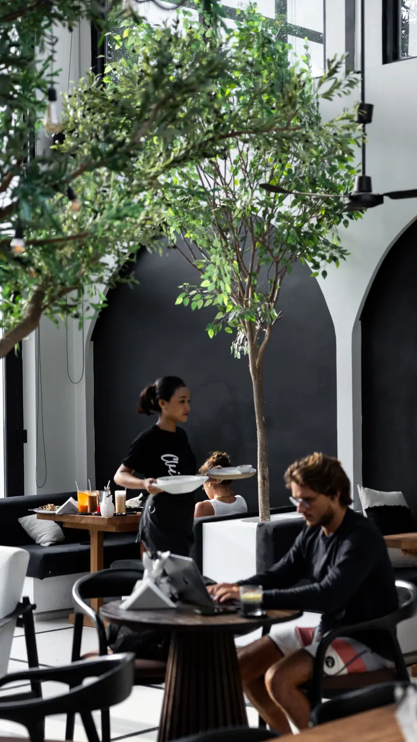 The high white room with black arches and trees. A waitress brings plates.