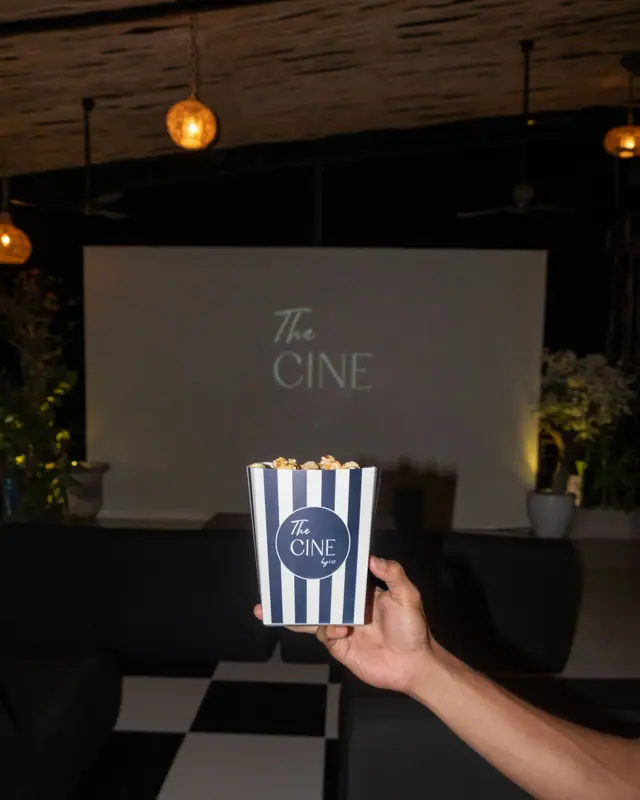Tangan mengangkat kotak popcorn bergaris di depan layar bioskop.