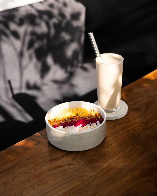 Smoothie bowl dan shake di sudut meja kayu, dalam cahaya yang menembus dedaunan.