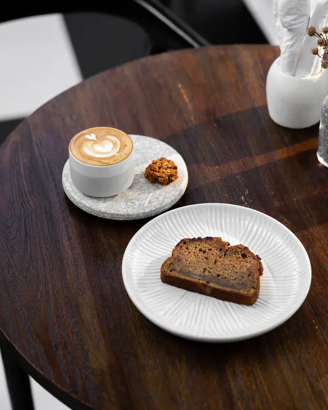 Flat white dan sepotong banana bread di meja kayu bundar.